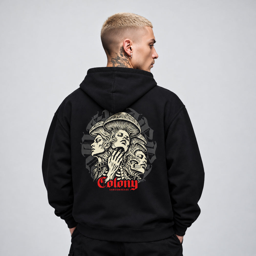 James wearing the black Colony hoodie, back view showing three interwoven mushroom-crowned portraits, a skull and studded collars, with red Colony lettering.