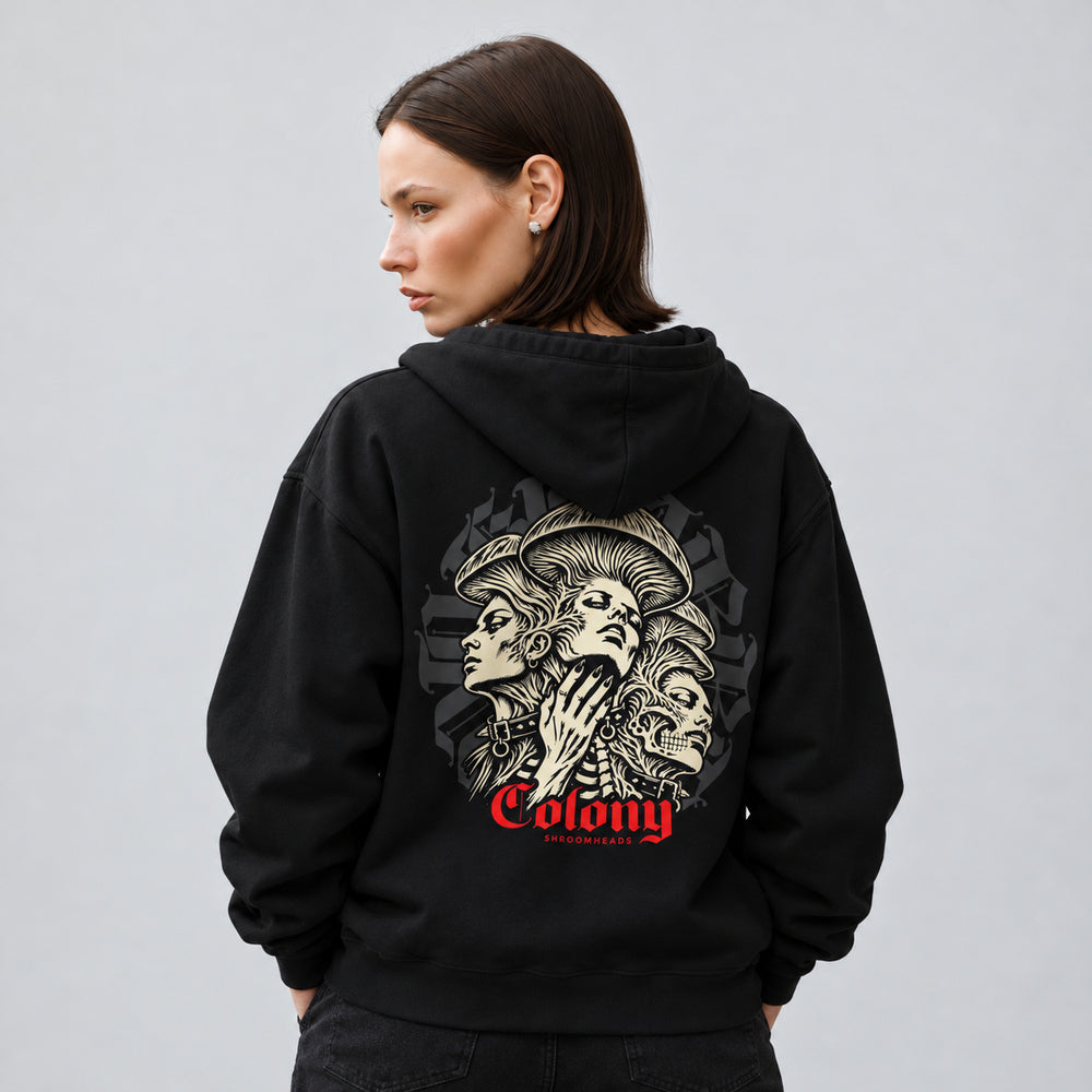 Mara wearing the black Colony hoodie, back view showing three interwoven mushroom-crowned portraits, a skull and studded collars, with red Colony lettering.