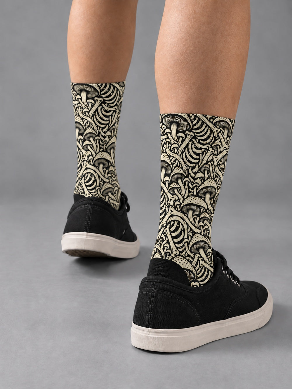 Rear three-quarter view of black crew socks with ivory mushroom and skeletal artwork, worn with black low-top canvas shoes.