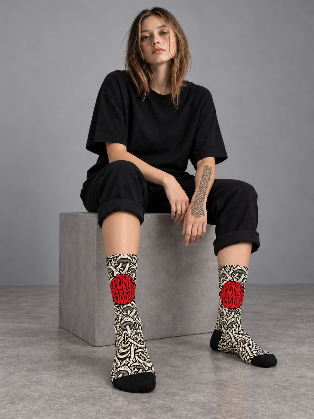 Mara seated in a gray studio wearing black crew socks with ivory mushroom and skeletal artwork and a red circular Punkrabbits logo, a plain black T-shirt and rolled black trousers.