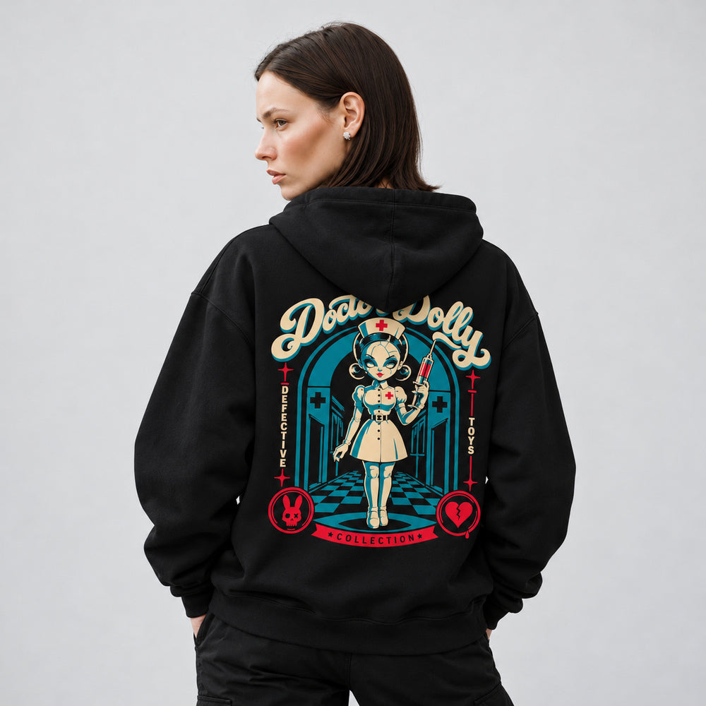 Mara wearing the black Doctor Dolly hoodie, back view showing a cracked porcelain nurse doll holding a red syringe in a teal hospital corridor, beneath cream Doctor Dolly lettering, with red rabbit and broken-heart emblems.