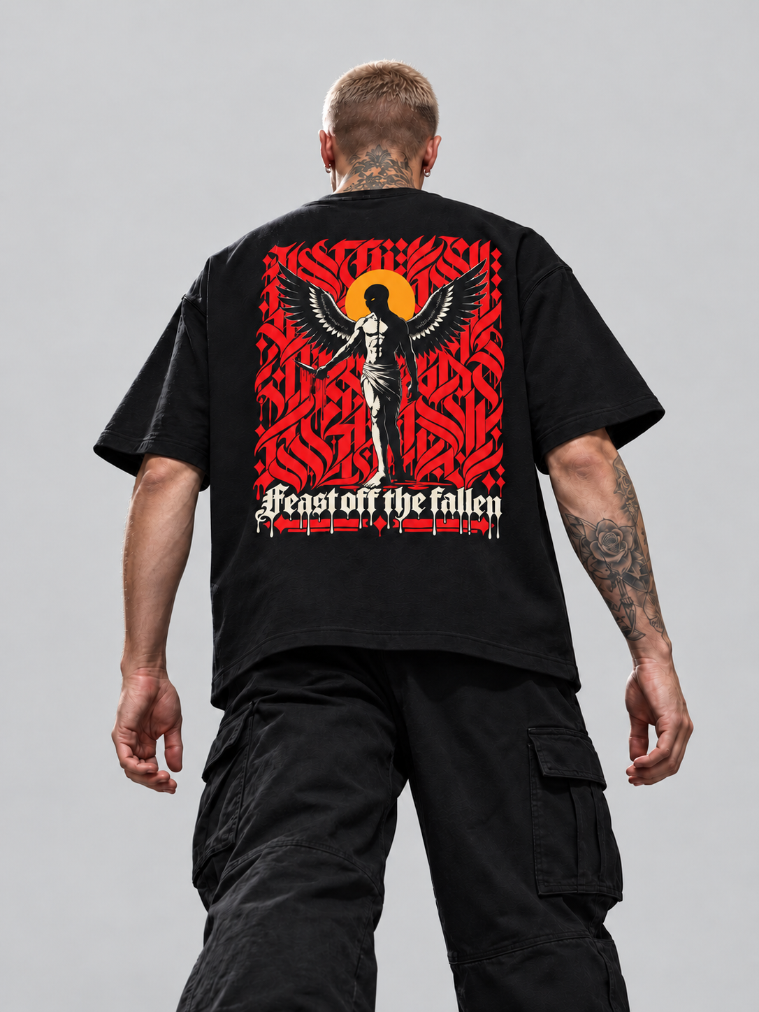 James wearing black Feast off the fallen Premium Boxy Tee, back view with winged figure, yellow-orange halo and red calligraphy in a gray studio.