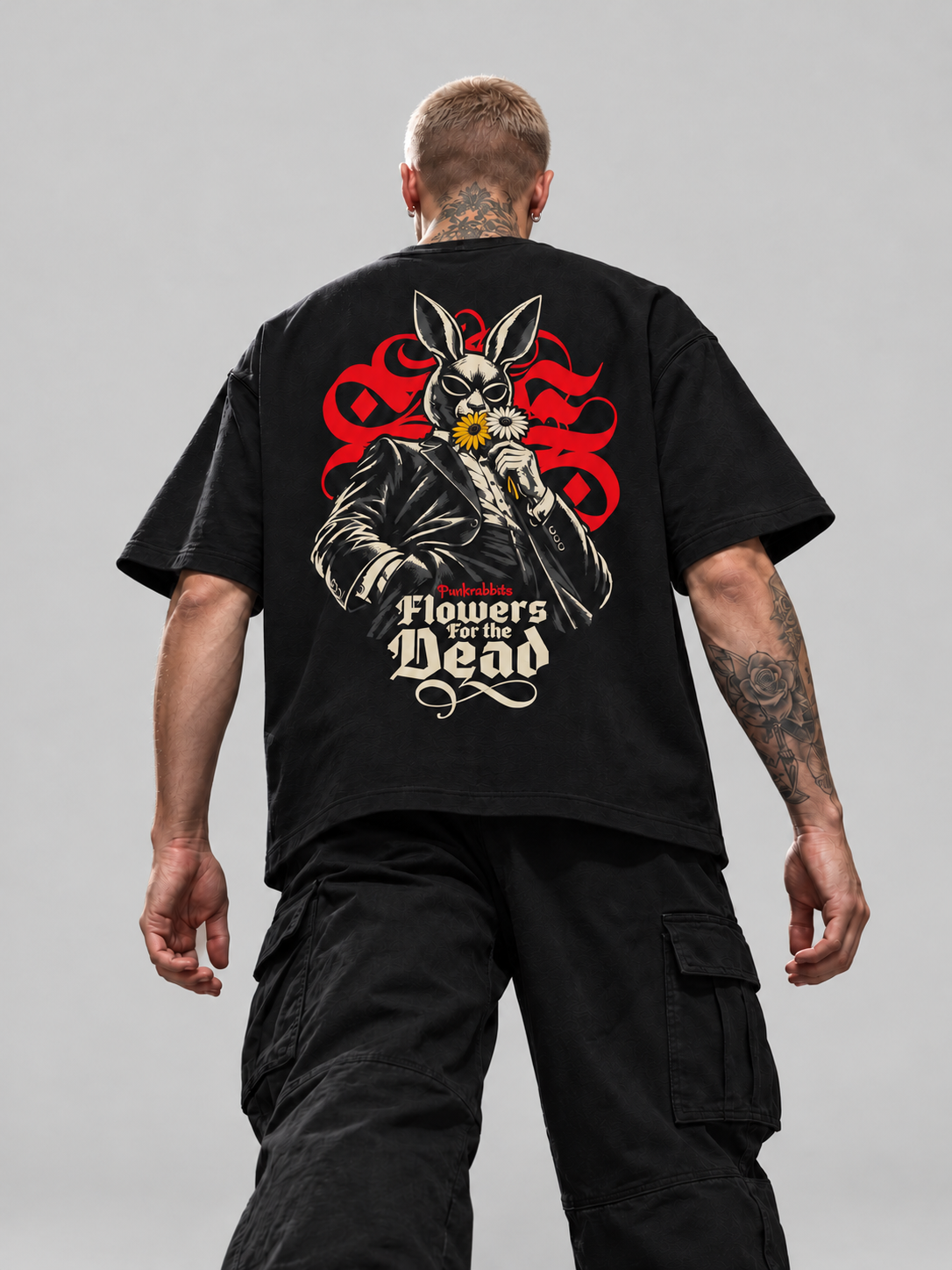 James wearing black Flowers for the dead Premium Boxy Tee, back view with suited rabbit-masked figure, flowers and red ornament in a gray studio.