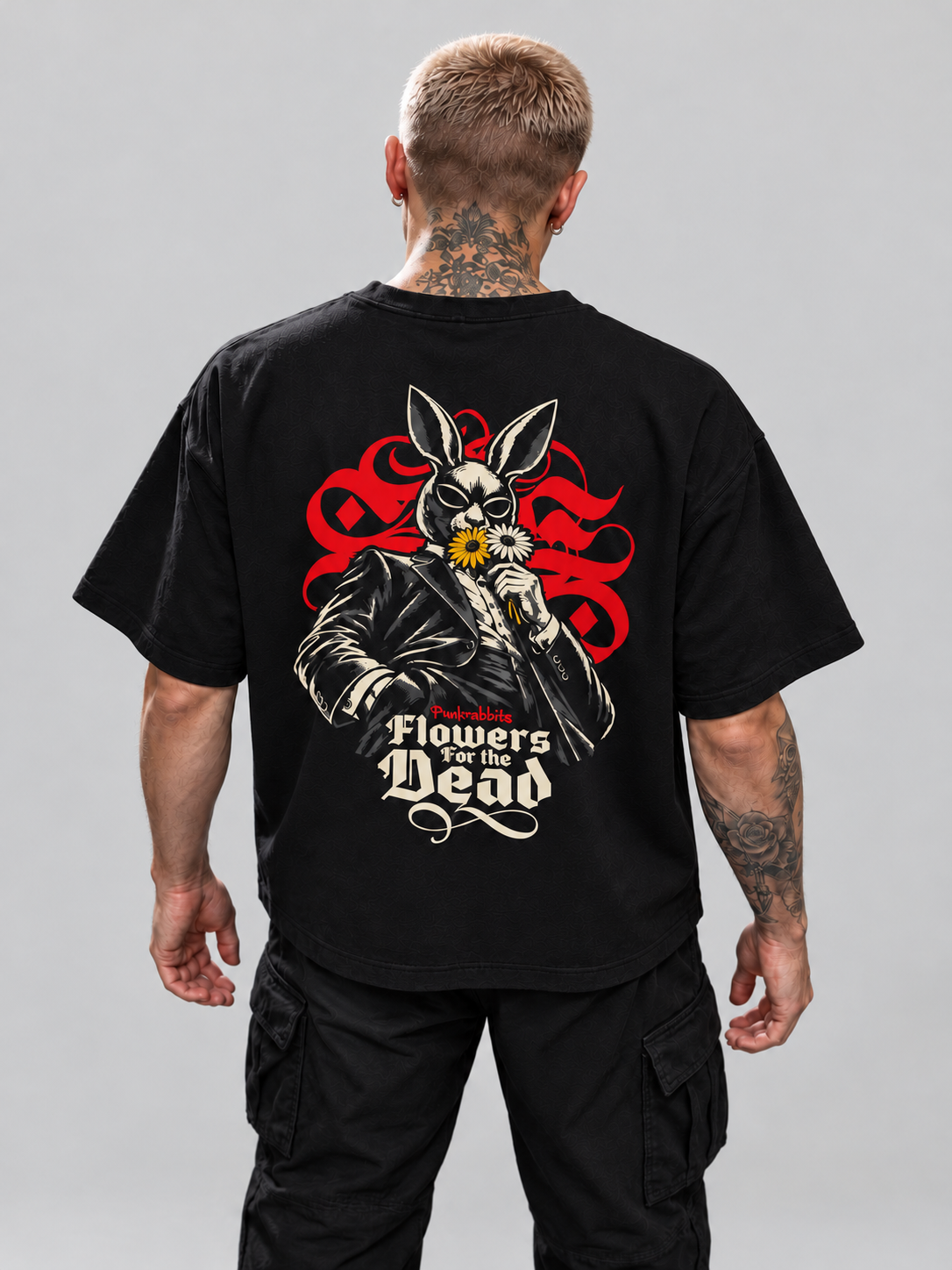 James wearing black Flowers for the dead Premium Boxy Tee, high-angle back view with suited rabbit figure and flowers in a gray studio.