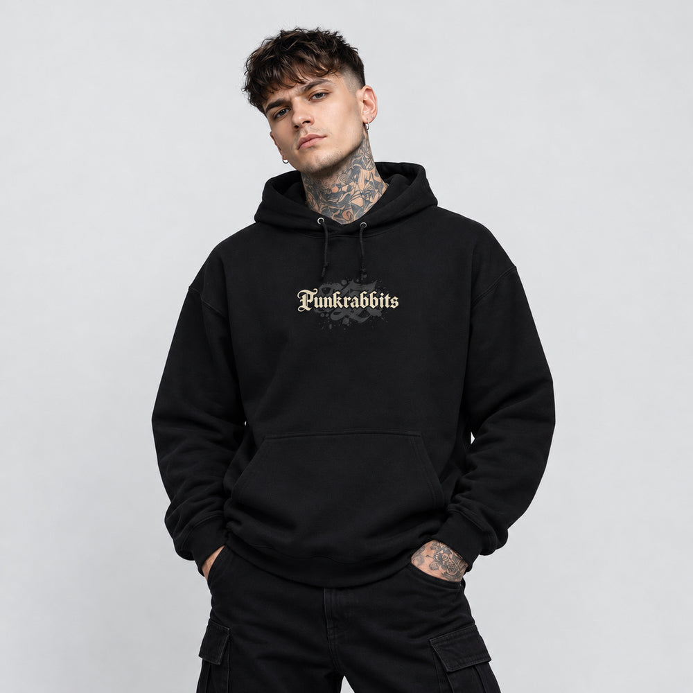 Noah wearing the black Hostile Takeover hoodie, front view showing cream Punkrabbits blackletter wordmark with a dark charcoal ornamental motif.