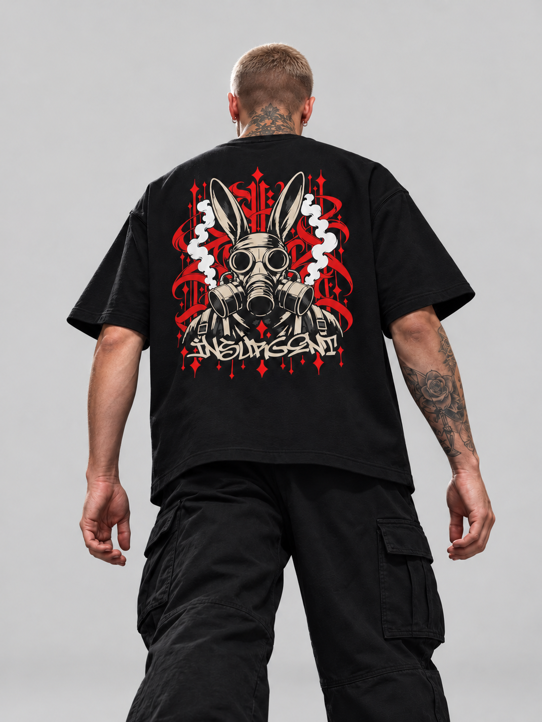 James wearing black Insurgent Premium Boxy Tee, low-angle back view with rabbit-eared gas mask and red calligraphy in a gray studio.