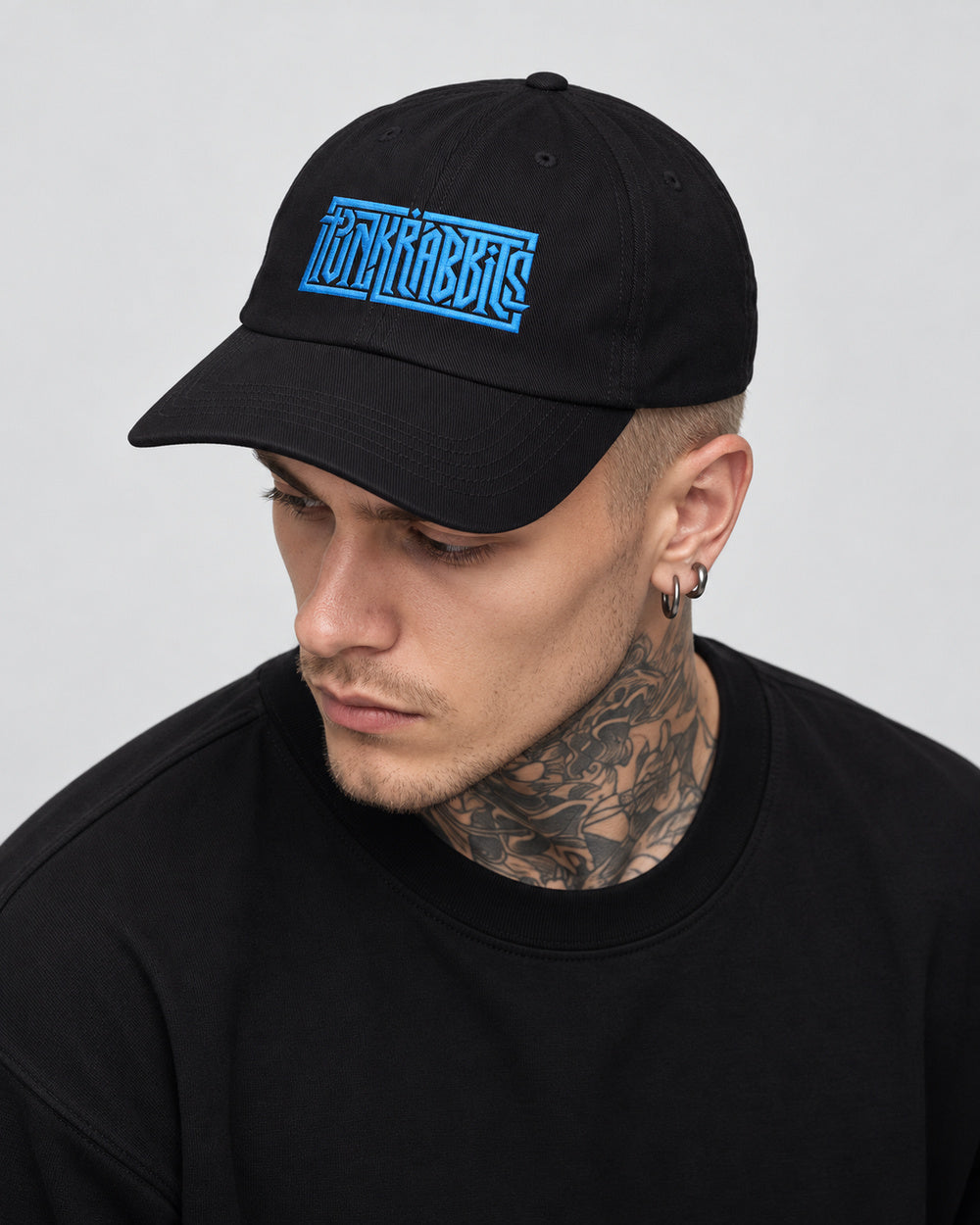 James wearing the black Punkrabbits cap with blue embroidery, looking down in a close gray-studio portrait.