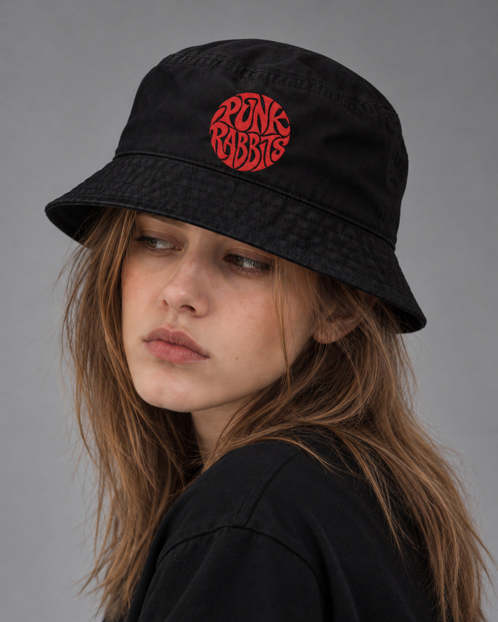 Mara turning away with her gaze off-camera in a black bucket hat with red circular Punk Rabbits embroidery.