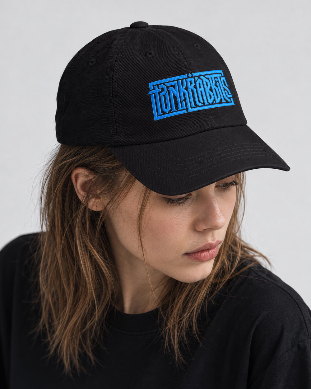 Mara wearing the black Punkrabbits cap with blue embroidery, looking down in a close gray-studio portrait.