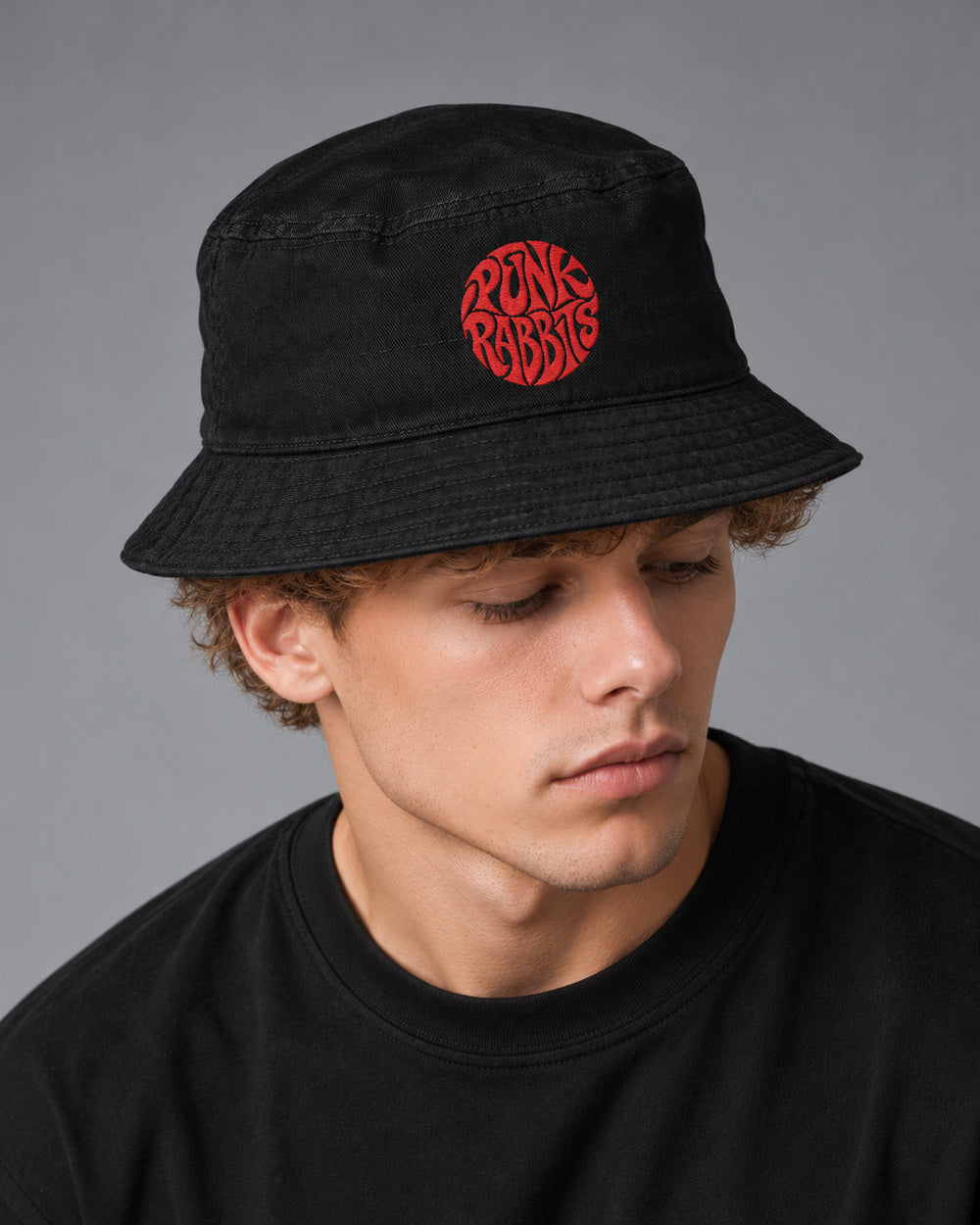 Theo looking down in a black cotton bucket hat with red Punk Rabbits embroidery, gray studio.