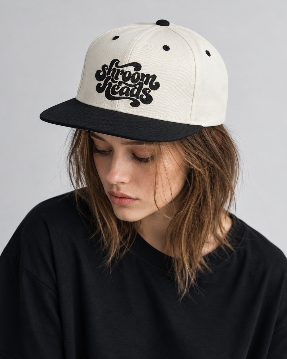 Mara wearing the Natural/Black Shroom Heads snapback, looking down in a gray studio portrait.