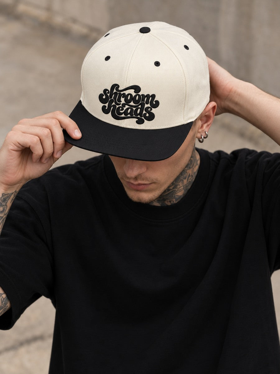 Mara wearing the Natural/Black Shroom Heads snapback, looking down in a gray studio portrait.