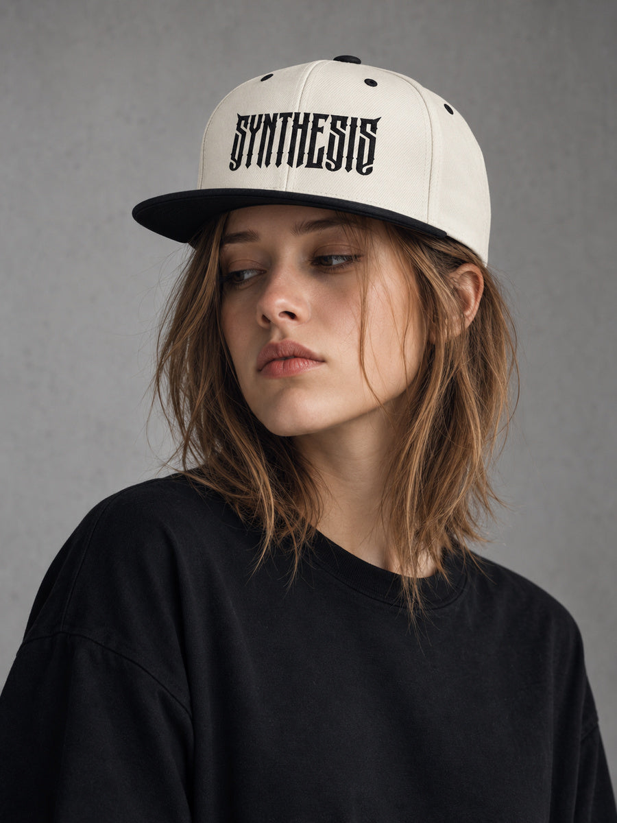 Mara looking away wearing the Synthesis Blackout snapback in Natural/Black, with black angular Synthesis embroidery and full flat bill visible.