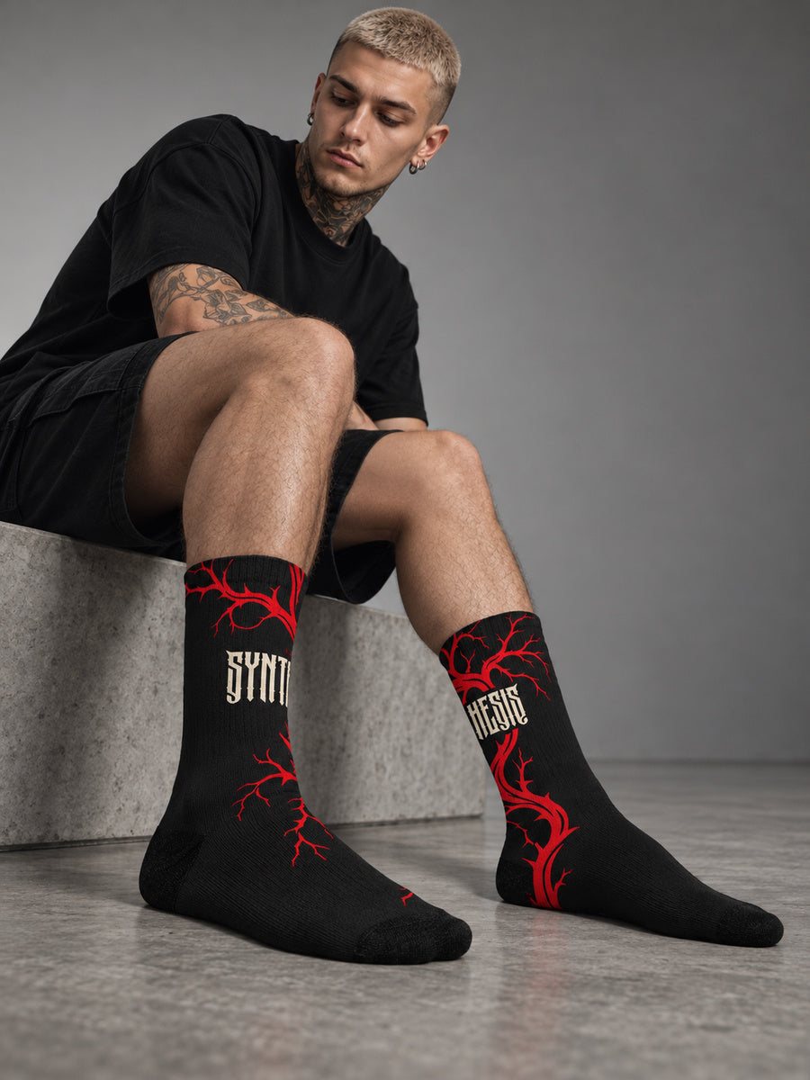 James seated in a gray studio wearing Bleeding Tree Synthesis crew socks, showing the opposite red branch and wrapped lettering sides with black toes and heels.