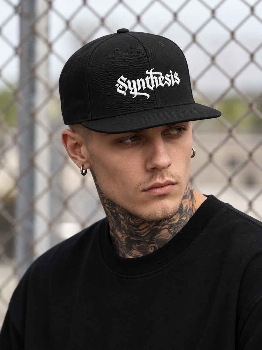 James wearing the Black Synthesis Doomed flat-bill snapback, seated low at the temples, looking away beside a chain-link fence.