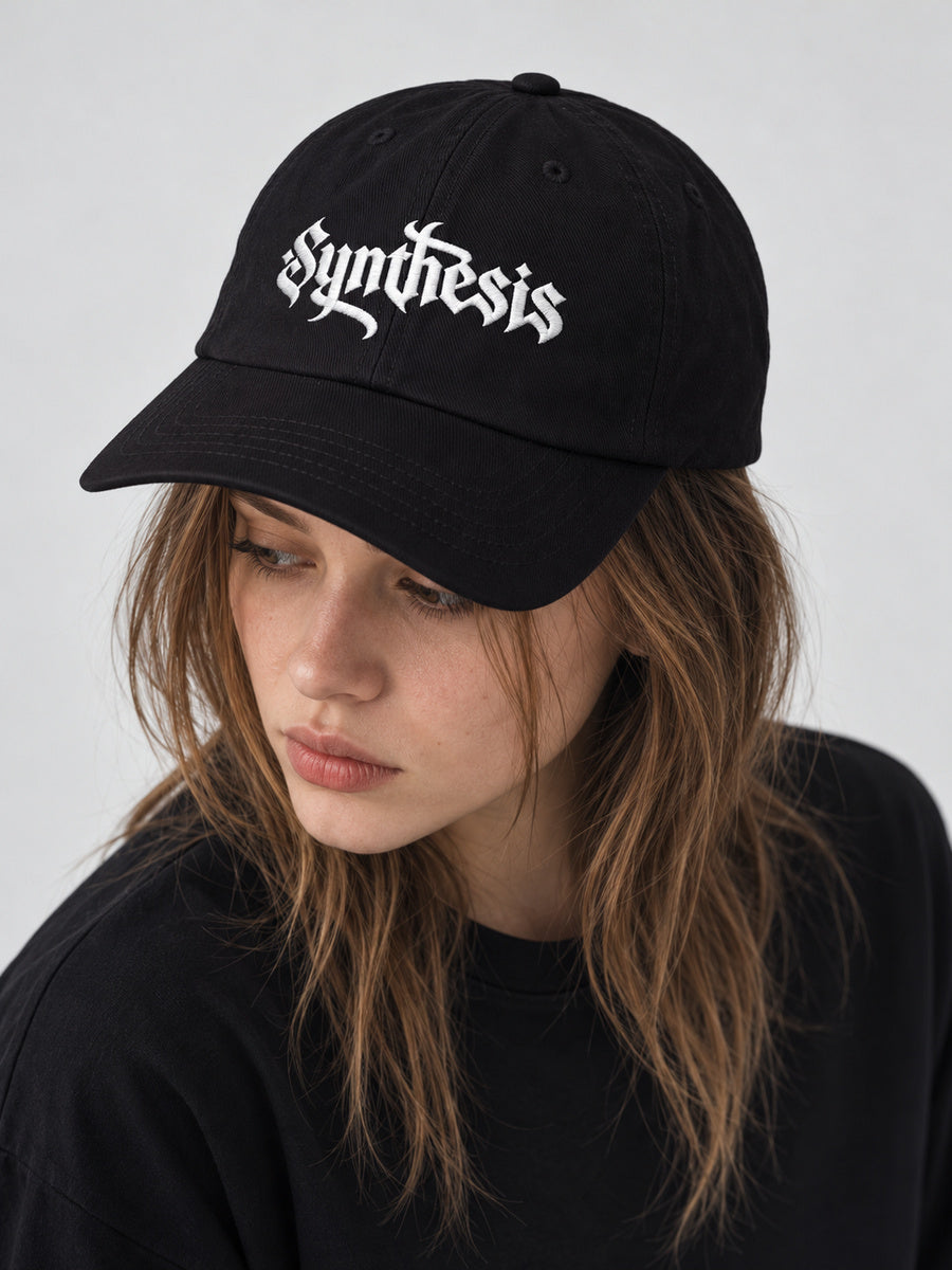 Mara wearing the black Synthesis Ritual cap, looking away in the gray studio.