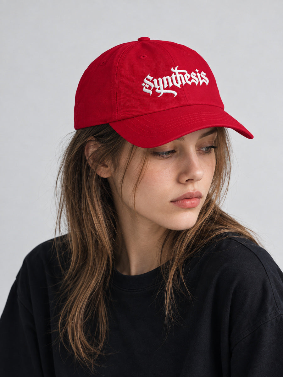 Mara wearing the cranberry Synthesis Ritual cap, looking away in the gray studio.