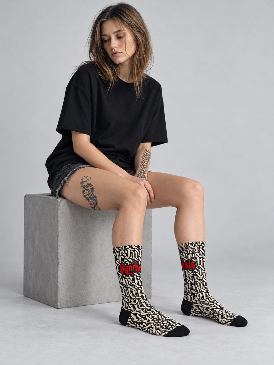 Synthesis script socks: Mara seated in the gray studio looking away, complete sock pair visible.