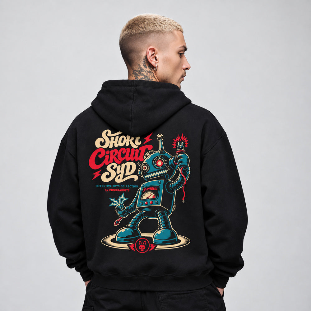 James wearing the black Short Circuit Syd hoodie, back view showing a teal retro toy robot with spring limbs, loose sparking cables, a chest meter and cream-and-red Short Circuit Syd lettering.