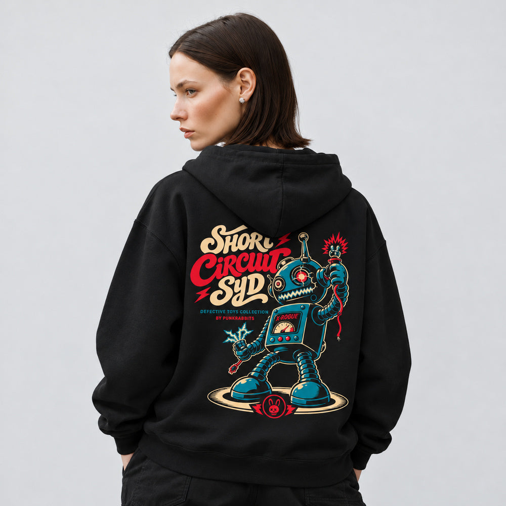 Mara wearing the black Short Circuit Syd hoodie, back view showing a teal retro toy robot with spring limbs, loose sparking cables, a chest meter and cream-and-red Short Circuit Syd lettering.