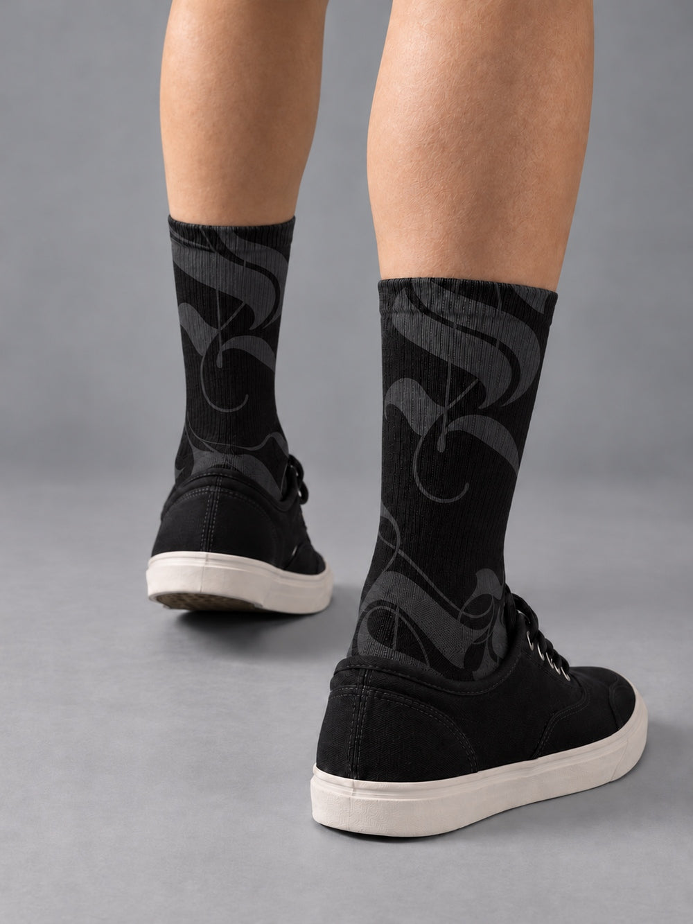 Rear three-quarter view of black crew socks with charcoal calligraphic strokes, worn with black low-top canvas shoes.