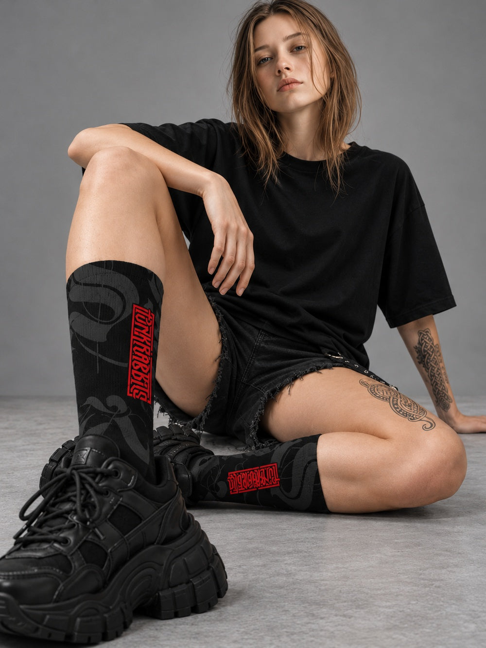 Mara seated in a gray studio wearing black crew socks with charcoal calligraphy and red Punkrabbits lettering, black shorts and chunky black shoes.