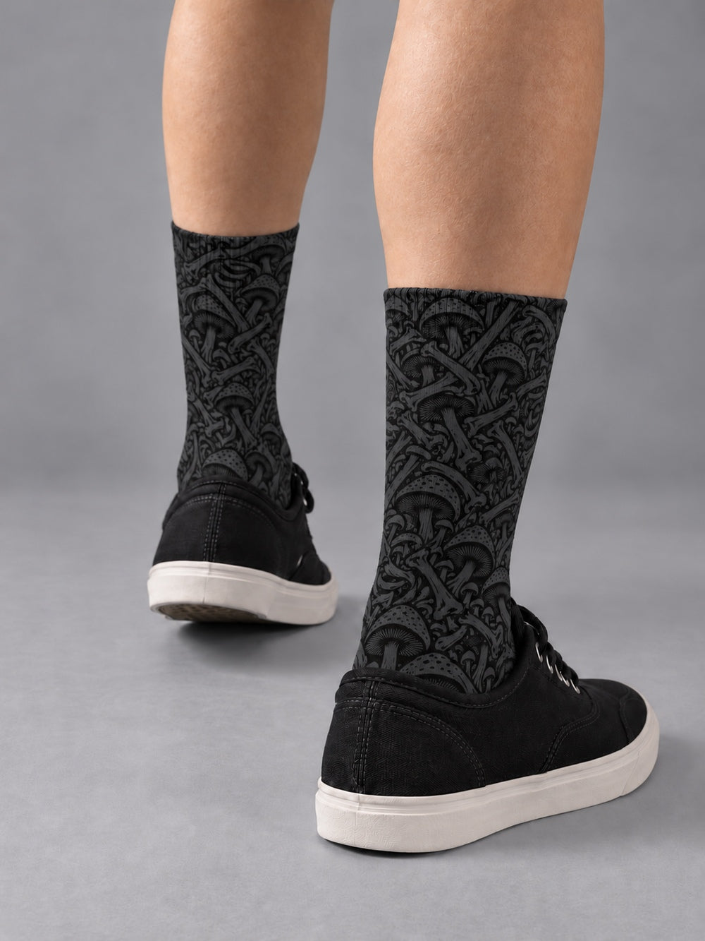 Rear three-quarter view of black crew socks with charcoal mushroom and skeletal artwork, worn with black low-top canvas shoes.