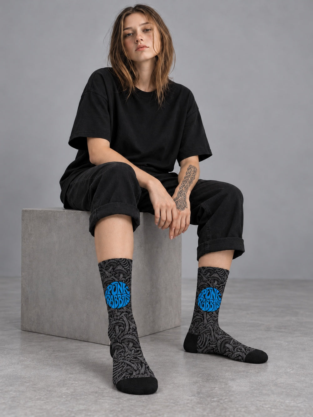 Mara seated in a gray studio wearing black crew socks with charcoal mushroom and skeletal artwork and a blue circular Punkrabbits logo, a plain black T-shirt and rolled black trousers.
