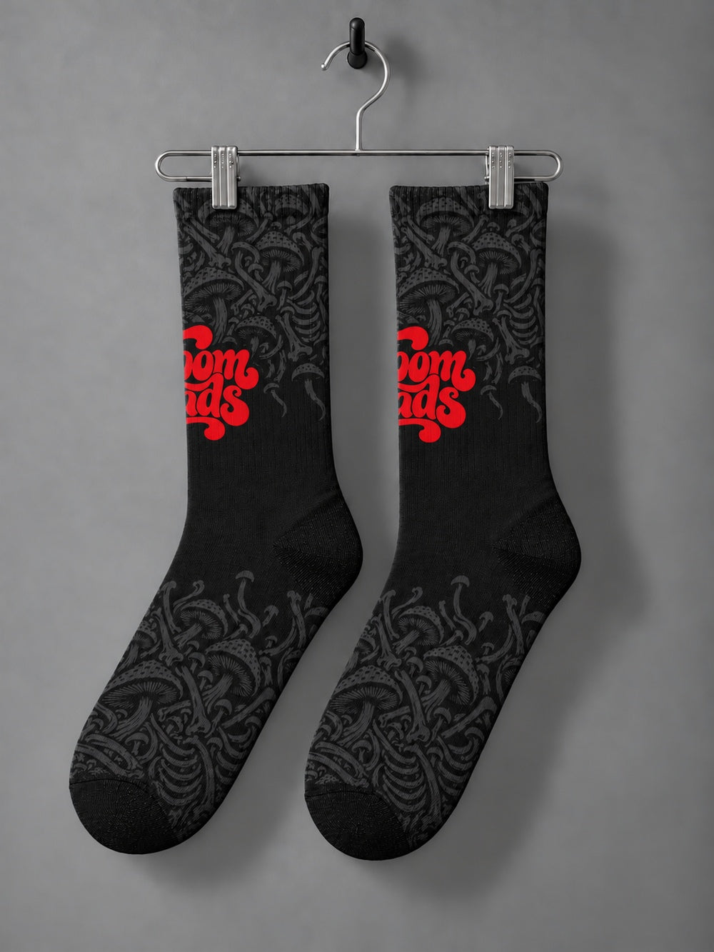 Pair hanging from a chrome clip hanger: black crew socks with fading charcoal mushroom artwork and red Shroomheads script.