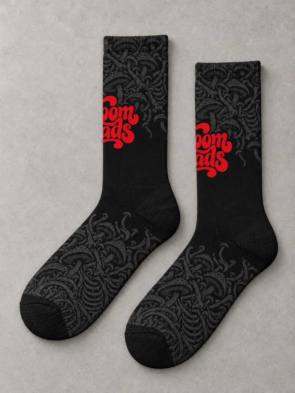 Overhead flat-lay pair in side profile: black crew socks with fading charcoal mushroom artwork and red Shroomheads script.