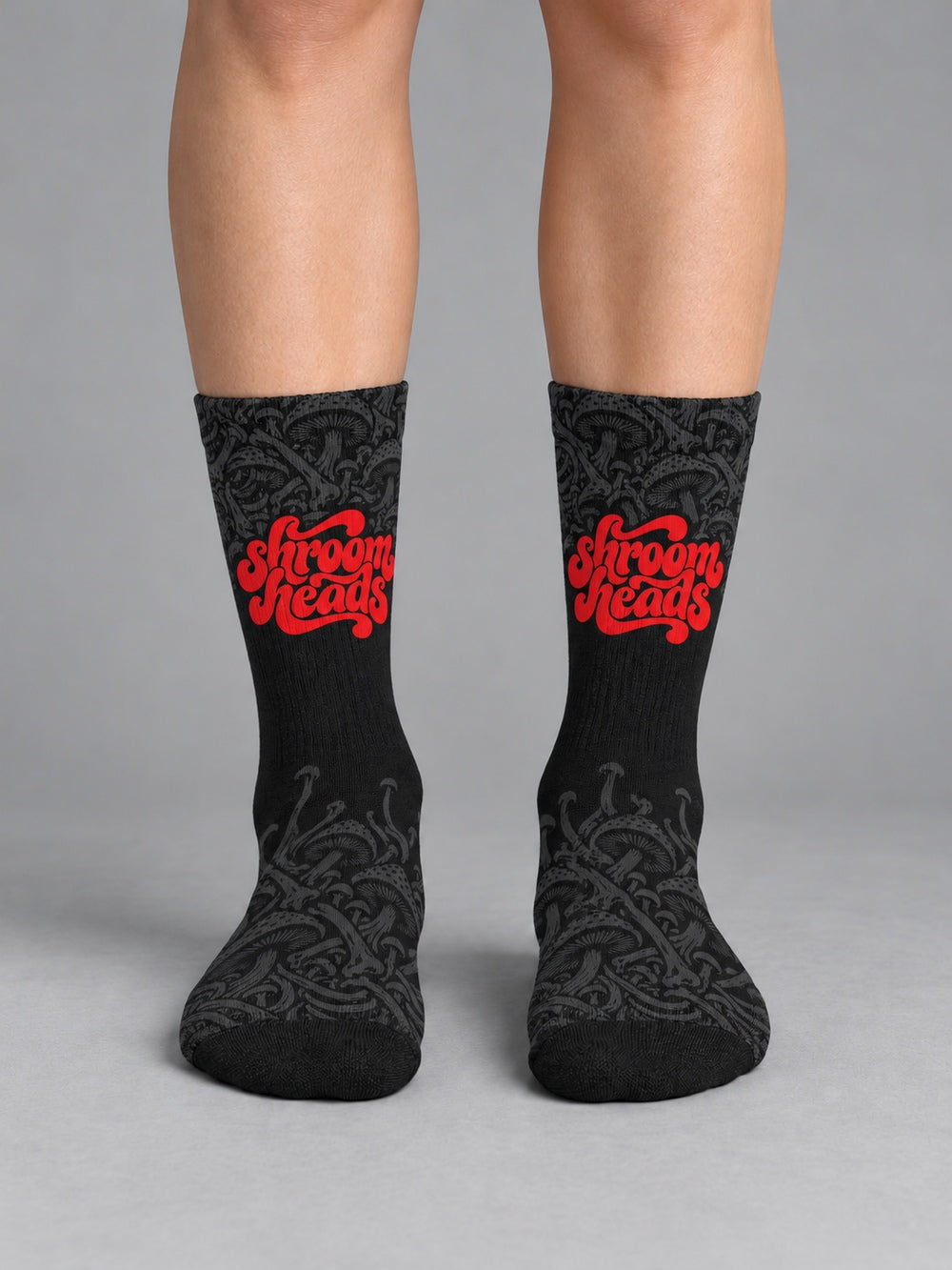 Front view worn on lower legs without shoes: black crew socks with fading charcoal mushroom artwork and red Shroomheads script.