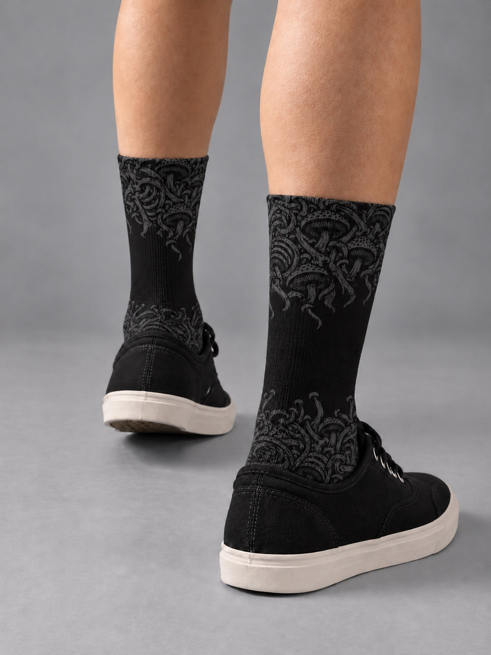 Rear three-quarter view of black crew socks with charcoal mushroom artwork fading into a black center, worn with black low-top canvas shoes.