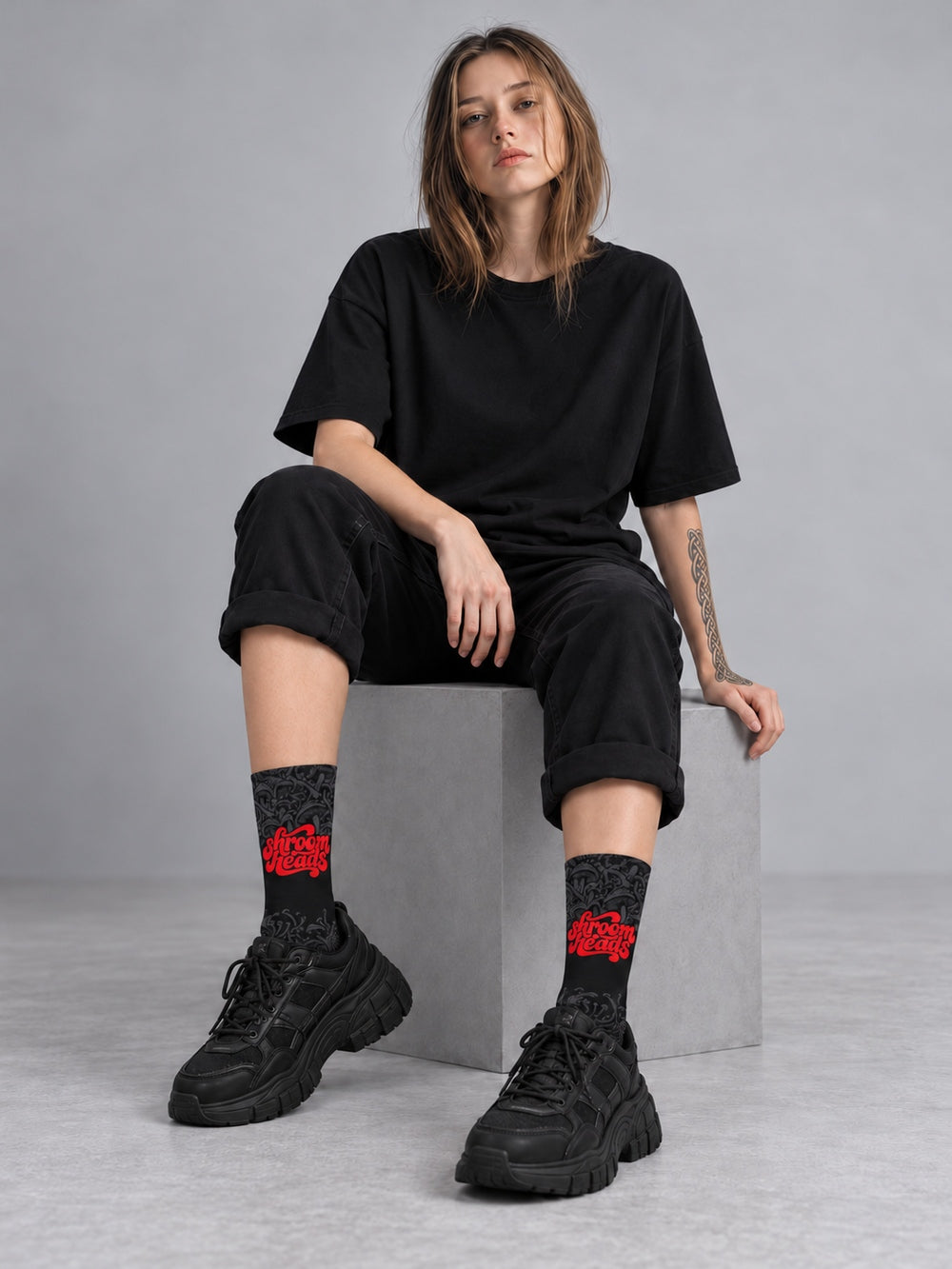 Mara seated in a gray studio wearing black crew socks with fading charcoal mushroom artwork and red Shroomheads script, a plain black T-shirt and rolled black trousers.