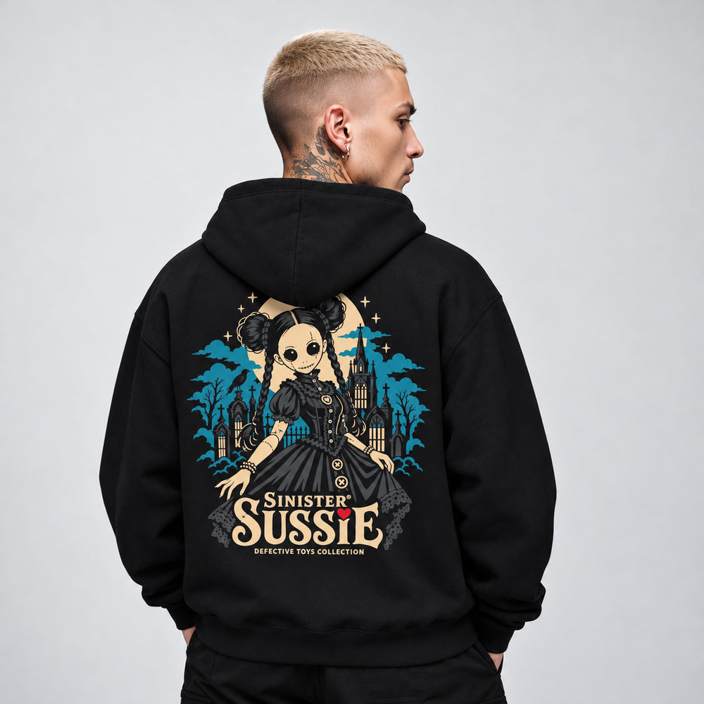 James wearing the black Sinister Sussie hoodie, back view showing a stitched doll with twin hair buns, braids and a charcoal dress before a teal Gothic church and cemetery, with an ivory moon, raven and Sinister Sussie lettering.