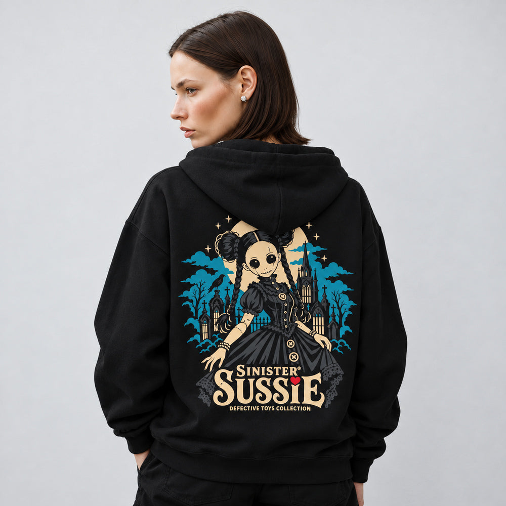 Mara wearing the black Sinister Sussie hoodie, back view showing a stitched doll with twin hair buns, braids and a charcoal dress before a teal Gothic church and cemetery, with an ivory moon, raven and Sinister Sussie lettering.