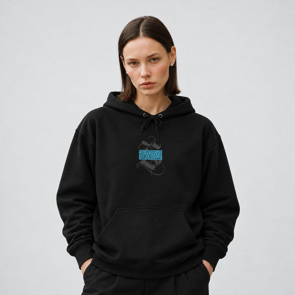 Mara wearing the black Spore Keeper hoodie, front view showing a compact blue Punkrabbits wordmark over a charcoal calligraphic motif.