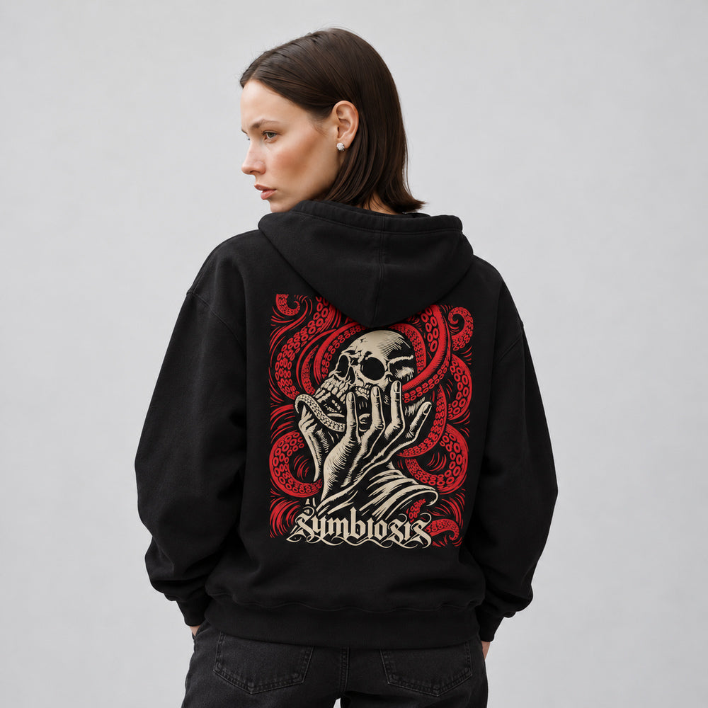 Mara wearing the black Symbiosis hoodie, back view showing an ivory skull cradled by an upward-reaching hand amid red octopus tentacles with suction cups, with ivory Symbiosis lettering.