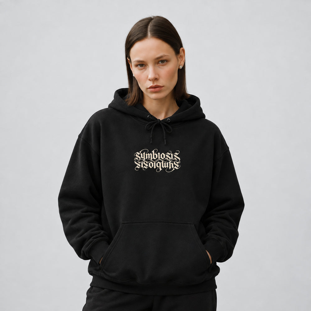 Mara wearing the black Symbiosis hoodie, front view showing paired upright and inverted ivory Symbiosis lettering.